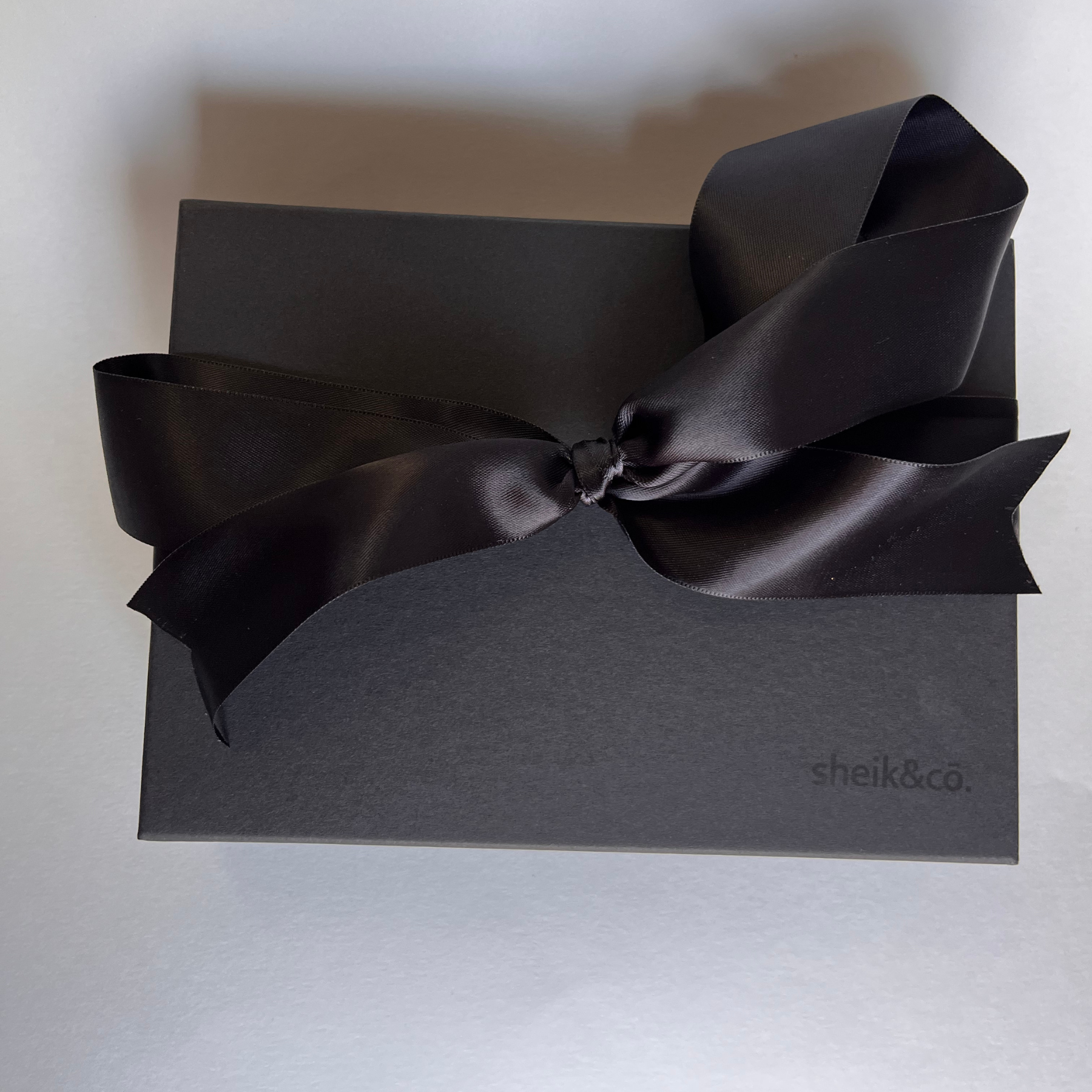 Black gift box with a large black ribbon on a white background, featuring the brand 'sheik&co'.